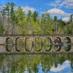 Stone arch bridge spanning a calm lake, with seven evenly spaced arches reflected almost perfectly in the still water. Tall pine and deciduous trees line the far shore beneath a bright blue sky with scattered clouds, creating a peaceful, mirror-like scene.