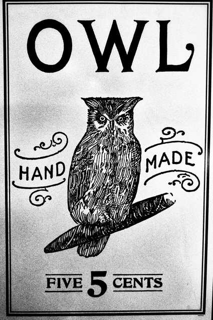 Black-and-white vintage-style sign featuring a detailed illustration of an owl perched on a rolled cigarillo. Large text at the top reads “OWL,” with decorative flourishes surrounding the words “HAND MADE.” At the bottom, the sign reads “FIVE 5 CENTS.” The design resembles an old apothecary, print shop, or market advertisement.