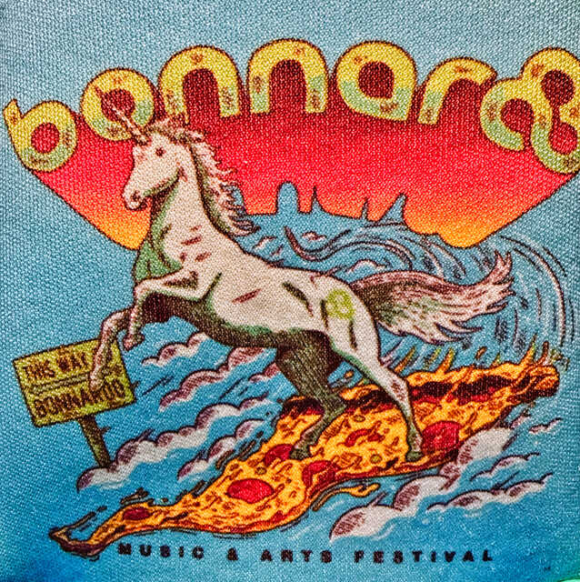Artwork for the Bonnaroo Music & Arts Festival. A white unicorn rears up while galloping across a giant slice of pepperoni pizza like a surfboard. A colorful sunset and stylized blue waves fill the background, with “bonnaroo” arched across the top in yellow letters. A small roadside sign reads “This Way → Bonnaroo.”
