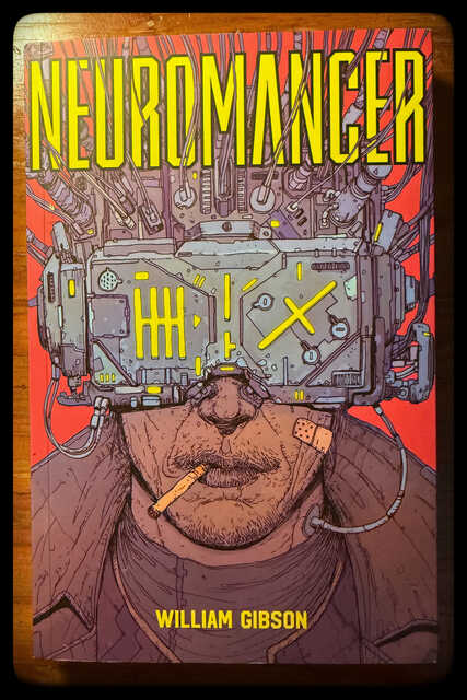 Book cover for Neuromancer by William Gibson. A man with a cigarette clenched between his lips wears a massive cybernetic visor that completely obscures his eyes. Thick cables and machinery connect the device to his head, while bright yellow lights glow across its surface. The background is vivid red, and the title appears in large yellow letters at the top. The overall style is gritty, cyberpunk, and industrial.