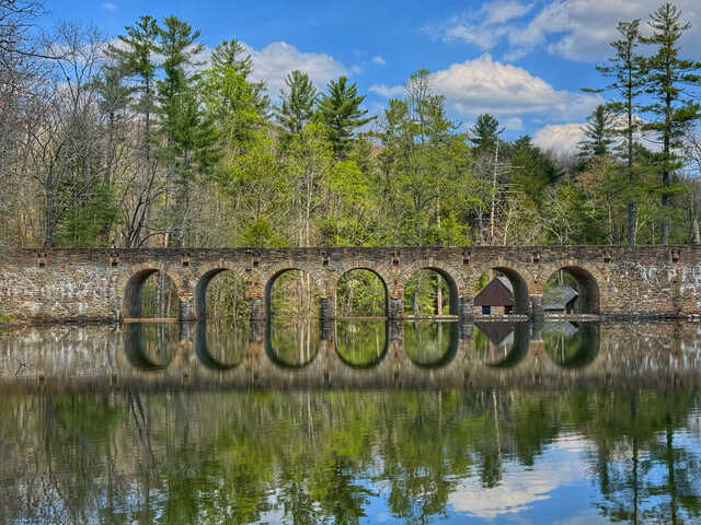 Stone arch bridge spanning a calm lake, with seven evenly spaced arches reflected almost perfectly in the still water. Tall pine and deciduous trees line the far shore beneath a bright blue sky with scattered clouds, creating a peaceful, mirror-like scene.